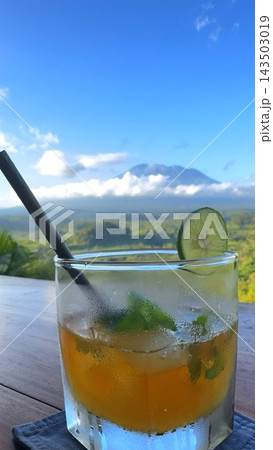 Iced cocktail rests on outdoor dining table, garnished with lime and mint beside straw. Condensation beads on glass as mountain and green valley complete this relaxed resort setting. 143503019