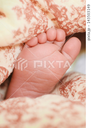 A tiny newborn foot peeks from warm patterned blankets in a soft macro closeup. The image reflects infant care, fragile life, parental tenderness, emotional comfort, and the first days at home. A tiny newborn foot peeks from warm patterned blankets in a soft macro closeup. The image reflects infant care, fragile life, parental tenderness, emotional comfort, and the first days at home. 143503149
