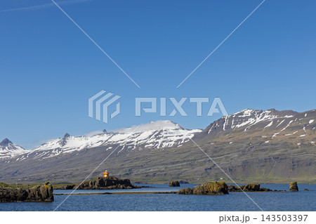 Orange lighthouse on rocky shore below snowy mountains in Eastfjords, Iceland 143503397