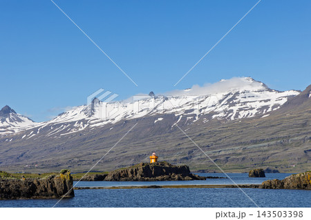 Orange lighthouse on rocky islet below snowy mountains in East Iceland 143503398