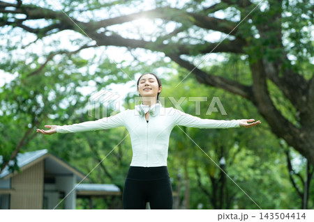Young Asian Woman Stretching Arms Open in Park with Eyes Closed, Feeling Free and Peaceful Outdoors Young Asian Woman Stretching Arms Open in Park with Eyes Closed, Feeling Free and Peaceful Outdoors 143504414