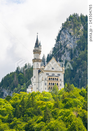 Neuschwanstein castle rises above green trees and rocky hills in Bavaria, Germany. Visitors can see the structure from a distance under a cloudy sky. Neuschwanstein castle rises above green trees and rocky hills in Bavaria, Germany. Visitors can see the structure from a distance under a cloudy sky. 143504765