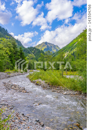 A river flows through the Weisspriach Valley in Austria, surrounded by green trees and high mountains under a blue sky with clouds. It is a bright day in the scenic landscape. A river flows through the Weisspriach Valley in Austria, surrounded by green trees and high mountains under a blue sky with clouds. It is a bright day in the scenic landscape. 143504766