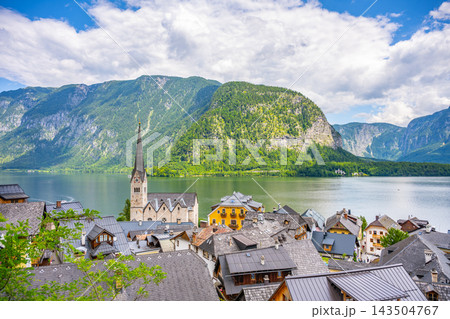 Hallstatt is a small town in Upper Austria with houses near the lake. A church tower stands tall among the rooftops. Mountains rise in the distance under a cloudy sky. Hallstatt is a small town in Upper Austria with houses near the lake. A church tower stands tall among the rooftops. Mountains rise in the distance under a cloudy sky. 143504767