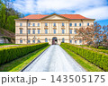 Boskovice Castle stands in Czechia with its classic Empire building style. The pathway leads to the front entrance, surrounded by trees and green bushes on a clear day. 143505175