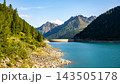 Lake Neves sits at the end of the Molini Valley in the Zillertal Alps, Italy. It features clear blue water and surrounding mountains with green trees. People may walk along the shore. 143505178