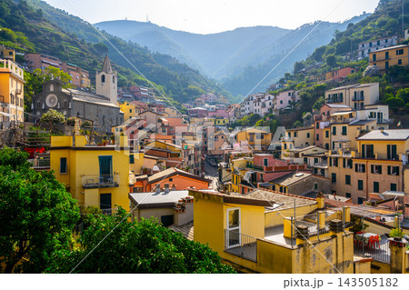 People enjoy the view of Riomaggiore village in Cinque Terre, Italy. The colorful buildings line the streets, surrounded by mountains and greenery. It is a bright day. People enjoy the view of Riomaggiore village in Cinque Terre, Italy. The colorful buildings line the streets, surrounded by mountains and greenery. It is a bright day. 143505182