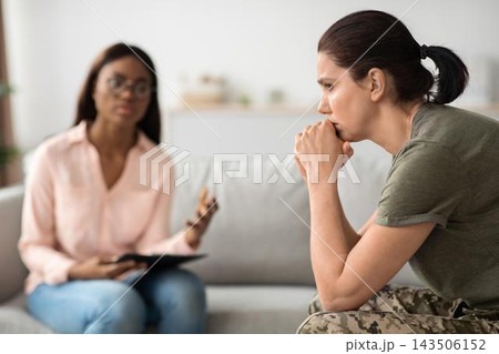 Worried Caucasian Female Veteran Deep In Thought During Therapy Session With Black Female Therapist In Office Setting, Addressing Emotional Health And Support Just Like Healthcare Concept. Worried Caucasian Female Veteran Deep In Thought During Therapy Session With Black Female Therapist In Office Setting, Addressing Emotional Health And Support Just Like Healthcare Concept. 143506152