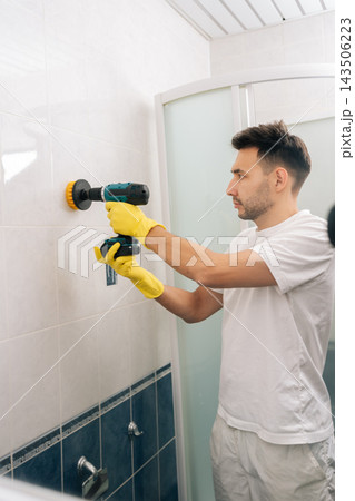 Vertical portrait of bearded young man wearing yellow gloves cleaning bathroom tiles using power drill with attached scrubber brush, streamlining household chores and deep cleaning surfaces. Vertical portrait of bearded young man wearing yellow gloves cleaning bathroom tiles using power drill with attached scrubber brush, streamlining household chores and deep cleaning surfaces. 143506223