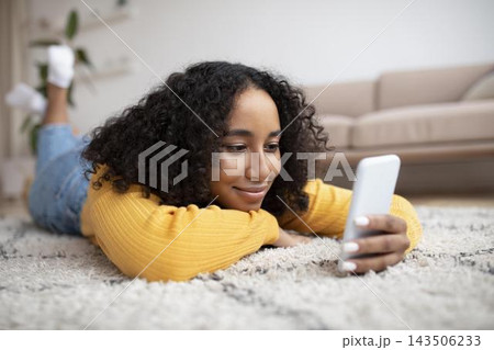 Smiling Black Young Woman Enjoying Time While Lying On Floor Using Smartphone At Home In Cozy Living Room, Engaging With Content On Screen, Relaxation Concept. Smiling Black Young Woman Enjoying Time While Lying On Floor Using Smartphone At Home In Cozy Living Room, Engaging With Content On Screen, Relaxation Concept. 143506233