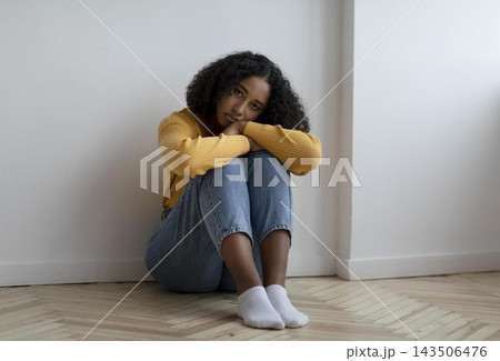 Worried Young Black Woman Sitting On Floor With Knees Hugged Close To Her Chest In A Minimalist Indoor Space. Reflecting Deep Thoughts In A Moment Of Introspection. Mental Health Concept. Worried Young Black Woman Sitting On Floor With Knees Hugged Close To Her Chest In A Minimalist Indoor Space. Reflecting Deep Thoughts In A Moment Of Introspection. Mental Health Concept. 143506476
