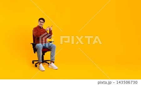 Smiling Young Asian Man Sitting In Office Chair And Pointing At Right, Expressing Confidence And Joy In Studio With Bright Yellow Background. Business Concept. 143506730