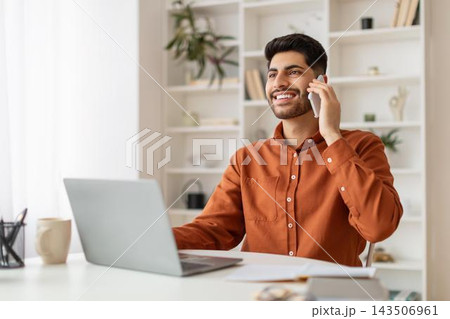 Smiling Young Man Of Middle Eastern Descent Talking On Phone While Sitting At Desk With Laptop, Engaged In Professional Work At Stylish Office, Reflecting Positive Business Concept. Smiling Young Man Of Middle Eastern Descent Talking On Phone While Sitting At Desk With Laptop, Engaged In Professional Work At Stylish Office, Reflecting Positive Business Concept. 143506961