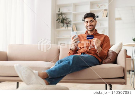 Happy Young Man Holding Credit Card And Smartphone While Relaxing At Home, Engaged In Online Shopping Experience. Sitting Comfortably On Sofa In Cozy Living Room, Enjoying Convenient Shopping. Happy Young Man Holding Credit Card And Smartphone While Relaxing At Home, Engaged In Online Shopping Experience. Sitting Comfortably On Sofa In Cozy Living Room, Enjoying Convenient Shopping. 143507085