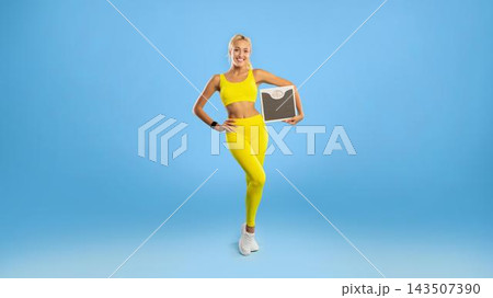 Smiling Young Woman In Yellow Activewear Holding A Weight Scale At Gym, Showcasing A Positive Emotion. Engaging In Fitness Routine In A Bright Studio Setting. Weight Loss Concept. 143507390