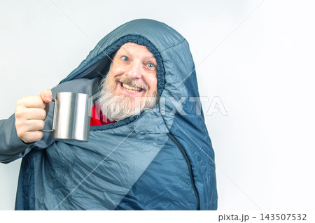 Man in a sleeping bag holds a mug and smiles in a bright indoor setting 143507532
