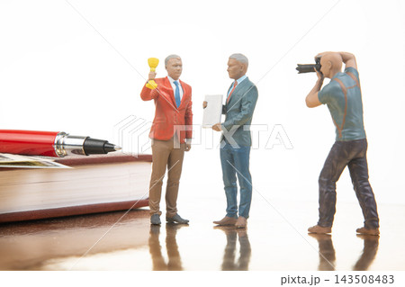 Miniature figures celebrate an achievement with a trophy and a camera in a simple office setting 143508483