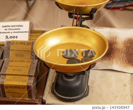 GANDIA, SPAIN - OCTOBER 04, 2026: Vintage mechanical balance scale with weights and artisan chocolate soaps displayed at a traditional outdoor market. GANDIA, SPAIN - OCTOBER 04, 2026: Vintage mechanical balance scale with weights and artisan chocolate soaps displayed at a traditional outdoor market. 143508947