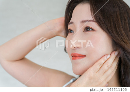 髪と頬に手を添えて優しく微笑む30代女性。美しいフェイスラインと透明感のある肌の美容イメージ 143511196