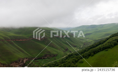 Lush green mountain valley with winding road under overcast sky and low morning mist Lush green mountain valley with winding road under overcast sky and low morning mist 143515736