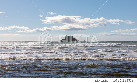 Ocean waves crashing near a rocky sea stack offshore under a cloudy blue sky Ocean waves crashing near a rocky sea stack offshore under a cloudy blue sky 143515805