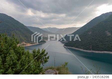 Scenic view of tranquil mountain lake surrounded by green hills under cloudy sky Scenic view of tranquil mountain lake surrounded by green hills under cloudy sky 143516206