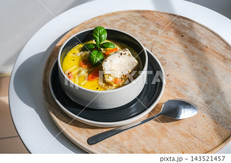 Sunlit Traditional Belgian Waterzooi Fish Soup. Sunlit bowl of traditional Belgian waterzooi fish soup on a wooden board, a creamy broth with white fish, carrots and potatoes topped with basil, a rust 143521457
