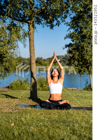 Young Woman in Seated Pose With Prayer Hands Overhead in a Green Park. A young Hispanic woman practises a seated pose with prayer hands overhead in a green park beside a calm lake at sunset, a moment Young Woman in Seated Pose With Prayer Hands Overhead in a Green Park. A young Hispanic woman practises a seated pose with prayer hands overhead in a green park beside a calm lake at sunset, a moment 143521951