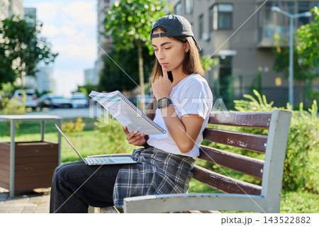 Young female student sitting on bench studying with notebook and laptop outdoors 143522882