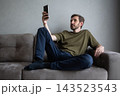 Young man relaxing on a comfortable sofa in his modern living room, holding a mobile phone and looking at the screen, engaging with digital content and enjoying casual leisure time 143523543