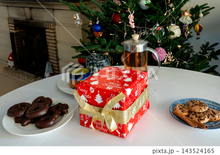 Red Christmas Gift French Press Tea and Cookies by Tree. A red Merry Christmas gift, a glass French press of tea and plates of chocolate gingerbread and chip cookies with cinnamon on a white table bef 143524165