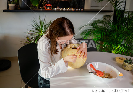 Daughter Reaching into Hollowed Pumpkin for Halloween Carving. A focused daughter with glasses reaches into a hollowed pale pumpkin to clean it at a white table with a bowl of seeds. A cozy autumn sce Daughter Reaching into Hollowed Pumpkin for Halloween Carving. A focused daughter with glasses reaches into a hollowed pale pumpkin to clean it at a white table with a bowl of seeds. A cozy autumn sce 143524166