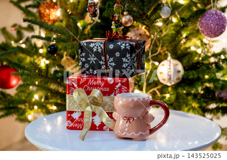 Gingerbread Man Mug with Stacked Christmas Gifts by Tree. A gingerbread-man mug sits beside stacked festive gifts, including a red Merry Christmas box, on a white table before a glowing Christmas tree Gingerbread Man Mug with Stacked Christmas Gifts by Tree. A gingerbread-man mug sits beside stacked festive gifts, including a red Merry Christmas box, on a white table before a glowing Christmas tree 143525025