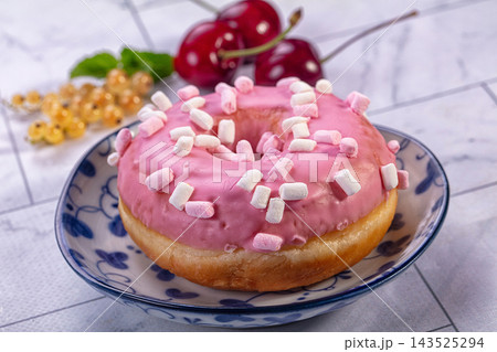 Pink glazed donut topped with mini marshmallows on blue patterned ceramic plate with fresh cherries and white currants in background, sweet pastry dessert 143525294