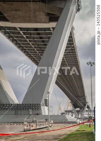 A low-angle view highlighting the imposing geometric architecture of a modern bridge superstructure, featuring a suspended work platform resting on the ground alongside safety cones, prepared for A low-angle view highlighting the imposing geometric architecture of a modern bridge superstructure, featuring a suspended work platform resting on the ground alongside safety cones, prepared for 143525508