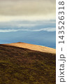 A sunlit mountain slope with dark shadows contrasts with a hazy valley and layered mountain ranges under a low cloud ceiling. 143526318