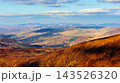 An aerial view of a mountainous landscape with rolling hills, distant wind turbines, and a blue sky with clouds. 143526320