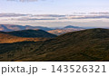 Golden afternoon light illuminates the vast grassy slopes and distant mountain ranges in Ireland as soft clouds drift across the horizon during the daytime. 143526321