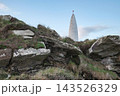 A tall, conical white structure with a red spherical top stands against a pale blue sky, partially obscured by rocky terrain. 143526329