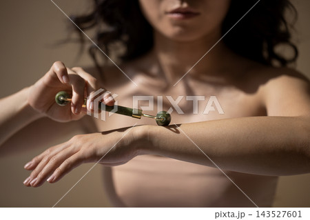 Woman applying a small metal roller to forearm with focused skincare gesture and bare shoulders against a softly blurred neutral background, close up beauty treatment scene 143527601