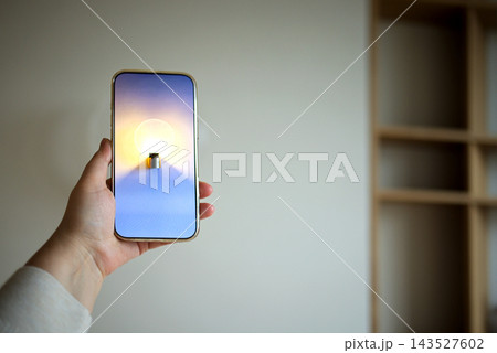 Smartphone mock up held in hand with blank gradient screen and glowing candlelike object, against softly lit wall and wooden shelving in an interior setting 143527602
