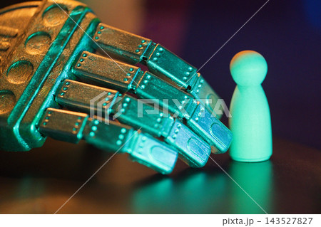 Robotic metallic hand reaching towards a small wooden figurine, illuminated with vibrant blue and green lighting against a dark background 143527827