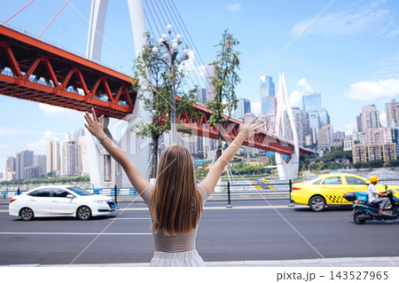 Young woman with long hair raises arms in excitement near a modern bridge and city skyline, with cars and trees visible along the road 143527965