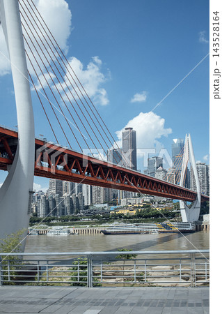 Modern cable-stayed bridge with red steel structure spans river, surrounded by urban skyline of high-rise buildings under a clear blue sky in Chongqing, China Modern cable-stayed bridge with red steel structure spans river, surrounded by urban skyline of high-rise buildings under a clear blue sky in Chongqing, China 143528164