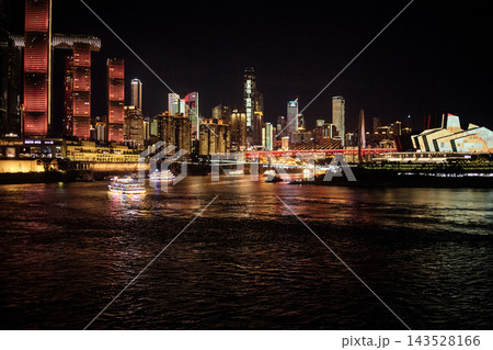 Nighttime cityscape of Chongqing, China featuring illuminated skyscrapers, boats on the river, and vibrant reflections on the water surface 143528166