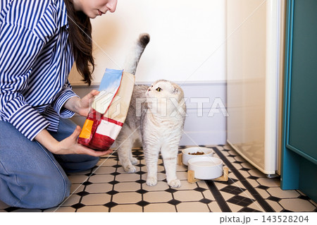 Woman in striped shirt kneeling on tiled floor holds cat food bag while interacting with a gray cat near food bowls in a modern kitchen setting Woman in striped shirt kneeling on tiled floor holds cat food bag while interacting with a gray cat near food bowls in a modern kitchen setting 143528204