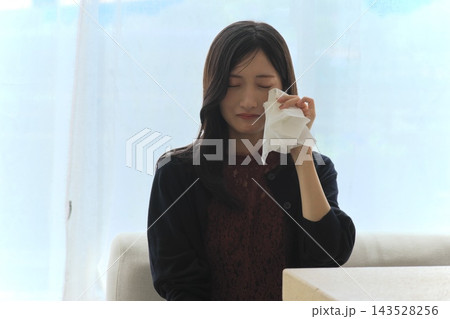 窓際でティッシュを手に目元の涙を拭い悲しむ若い女性（悲しい・涙・ショック・お悩みイメージ） 143528256