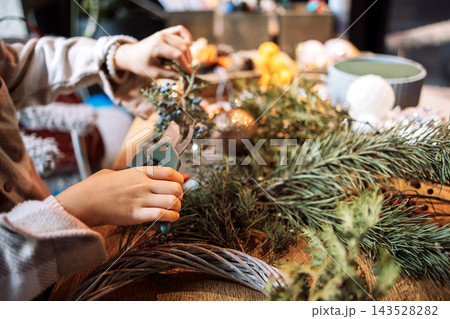 Child's hand using pruning shears to cut greenery for holiday wreath decoration on wooden table with various festive ornaments and materials visible 143528282