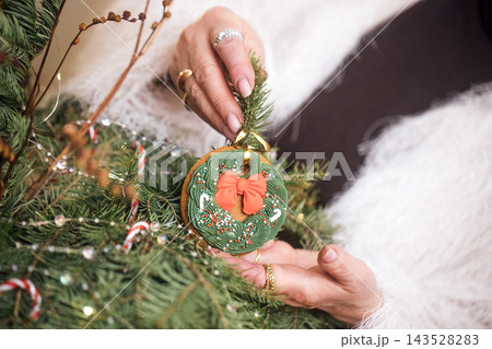 Woman's hands holding a decorative Christmas ornament with a red bow, surrounded by evergreen branches and festive decorations in a cozy indoor setting 143528283