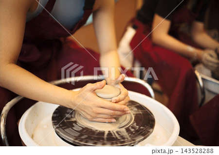 Female potter shaping clay on a pottery wheel in a workshop, with other artisans visible in the background working on their projects Female potter shaping clay on a pottery wheel in a workshop, with other artisans visible in the background working on their projects 143528288
